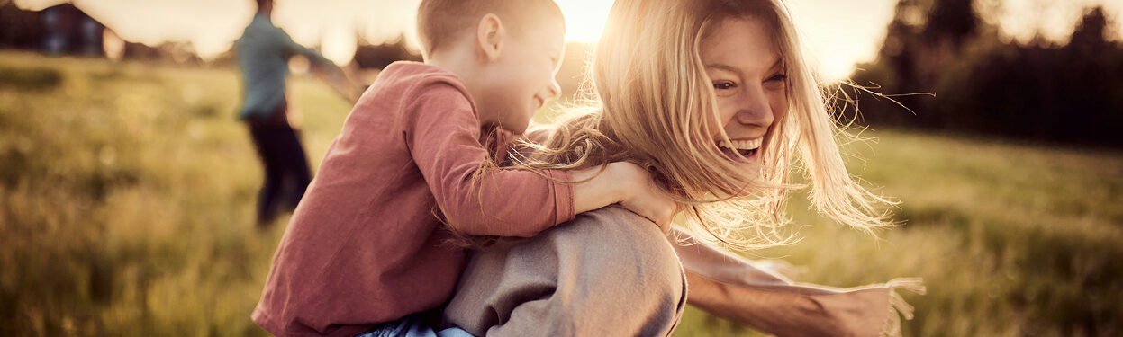 A mother holds her young son on her back for a piggyback ride. They're situated in a beautiful park with a newly built home development in the background.
