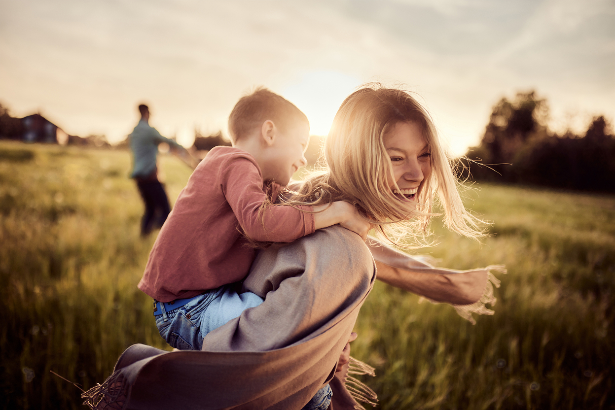 A mother holds her young son on her back for a piggyback ride. They're situated in a beautiful park with a newly built home development in the background.