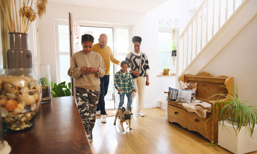 A young family enters their home through the front door. Father, mother and two young daughters. The youngest daughter leads the family dog on a lease.